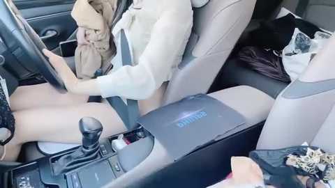 Media: Video of a person in a beige jacket, seated in a car, with a beige steering wheel, gear shift, and center console. The car's interior is light-colored, and there's a bag on the seat.