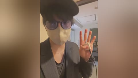 Media: Video of a person in a dark suit, wearing a black cap, black sunglasses, and a beige face mask, with red-painted nails, waving from behind a partially open door.