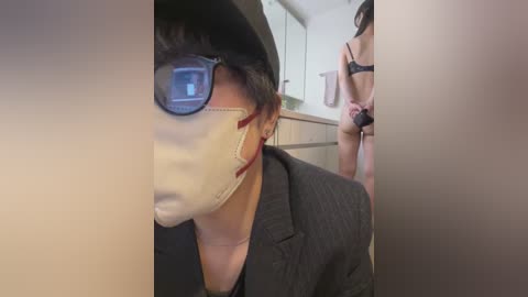 Video of a woman in a black pinstripe suit and white mask with glasses, standing beside a woman in a black bra and panties in a bathroom.