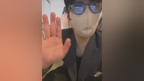 Media: Video of a person with light skin, wearing a black suit, white face mask, and dark sunglasses, holding up a hand against a blurry, possibly reflective background.