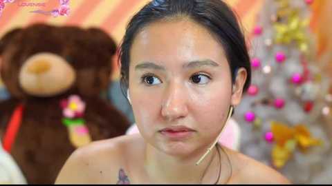 Media: Video of a young Asian woman with dark hair, light skin, and earrings, looking serious, with a stuffed bear and festive background.