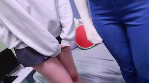 Media: Video showing a close-up of two people in casual attire: a person in a white sweater and grey shorts, holding a white bag with a red and green logo, standing beside a person in blue jeans, in a modern, indoor setting.