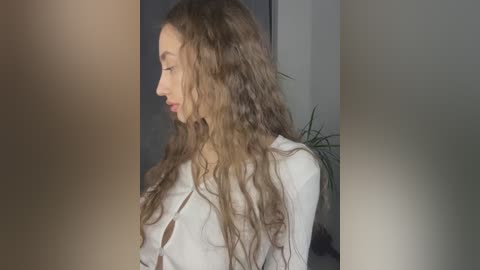 Media: Video of a fair-skinned woman with long, wavy, light brown hair, wearing a white blouse, seen from the side, in a dimly lit indoor setting.