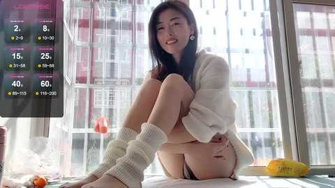 Media: A video of an Asian woman with long black hair, sitting on a bed in a modern room, wearing a white knit sweater and white leg warmers, with a digital clock and a banana on the bedside table.