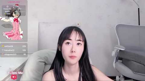 Media: A video of a young East Asian woman with long black hair and fair skin, wearing a pink dress, sitting in a modern living room with a white wall and grey furniture.