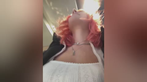 Media: Video of a woman with pink hair, wearing a white, lace-trimmed top, black jacket, and silver necklace, standing in a dimly lit room with blurred background.