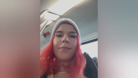 Media: A video of a young woman with light skin, pink hair, and a white beanie, smiling, taken from a low angle. She's in a bus, with a fluorescent ceiling light and a window in the background.