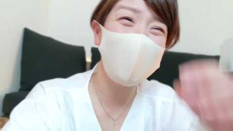 Media: Video of an East Asian woman with short brown hair, wearing a white mask and white robe, smiling with closed eyes. Background features dark cushions and a beige wall.