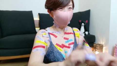 Media: Video of a young Asian woman with short brown hair, wearing a blue lace bra, with various colorful bandages on her face and chest, sitting in a modern living room with a dark couch and a small potted plant.