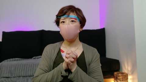 Media: Video of an Asian woman with short brown hair, wearing a green cardigan, light pink face mask, and blue headband, clasping hands in prayer position, in a modern living room with black sofa, gray blanket, and purple lighting.