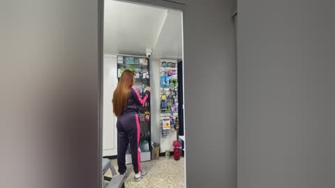 Media: Video of a woman with long, straight red hair in a black and pink tracksuit, shopping in a well-lit convenience store with shelves of snacks and drinks.