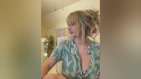 Media: Video of a blonde woman in a green floral blouse, sitting indoors, with a potted plant and beige curtains in the background.