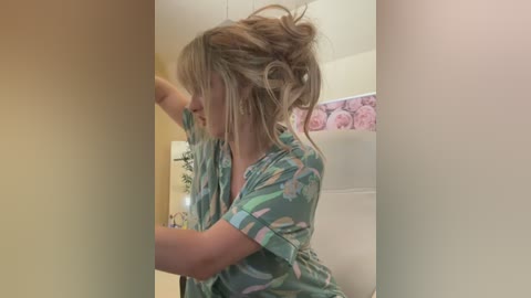 Media: Video of a woman with blonde hair styled in a messy updo, wearing a green floral-patterned shirt, standing in a bathroom with beige walls and floral wallpaper.