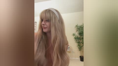 Media: A video of a blonde woman with long, wavy hair, wearing a white headband, standing in a brightly lit room with pale walls and a green plant.