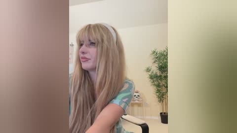 Media: Video of a blonde woman with long, straight hair and bangs, wearing a light blue top, standing in a room with a white wall, a green plant, and a black chair.