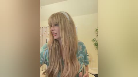 Media: Video of a blonde woman with long, wavy hair wearing a light blue floral top, standing in a bathroom with beige walls and a shower curtain.