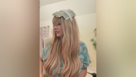 Media: Video of a woman with long, wavy blonde hair, wearing a light blue patterned shirt and a paper crown, standing in a pale yellow room with a leafy plant in the background.