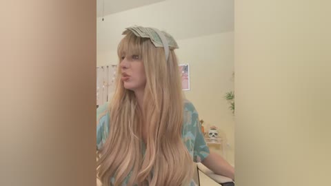 Media: Video of a young woman with long, straight blonde hair, wearing a teal and white patterned shirt, standing in a pale yellow room with a framed poster and plant in the background.