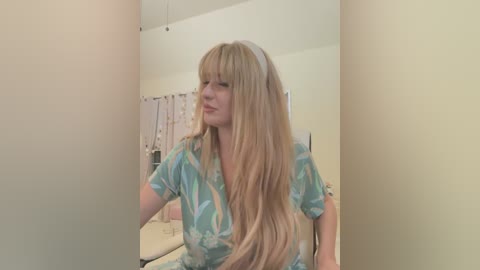 Media: Video of a fair-skinned woman with long, straight blonde hair in a tie-dye top, seated indoors in a minimalist room with cream walls and a white shower curtain.