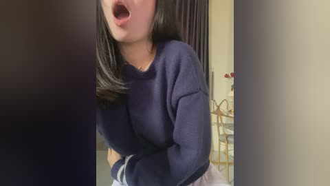 Media: Video of an Asian woman with shoulder-length black hair, wearing a navy blue sweater, mid-sentence, in a dimly lit room with a white table and dark curtains in the background.