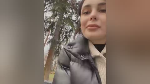 Media: A video of a young woman with light skin, dark hair, and brown eyes, wearing a black puffer jacket and beige sweater, standing outdoors with a blurred background of trees.