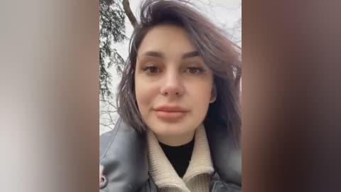 Media: Video of a young woman with fair skin, dark hair, and brown eyes, wearing a black top under a gray leather jacket, standing outdoors with a blurred background of leafless trees.