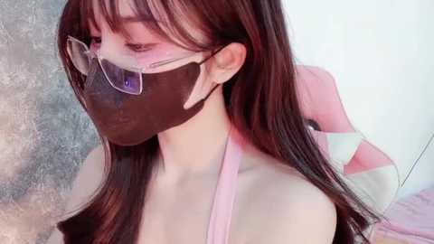 Media: Video of a young Asian woman with long, straight brown hair, wearing a black mask, glasses, and a pink top, standing against a textured, light-colored background.