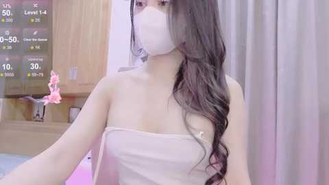 Media: Video of an Asian woman with long, wavy dark hair, wearing a white face mask, towel, and a pink bra, standing in a bathroom with wooden cabinets and a pink flower in the background.