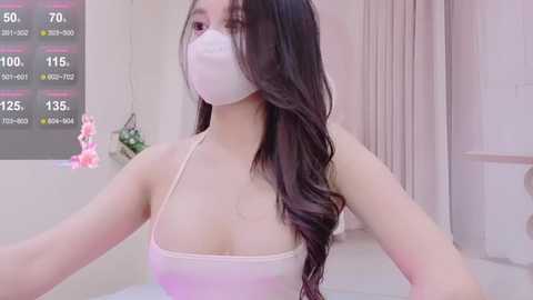 Media: Video of an Asian woman with long dark hair, wearing a white face mask, pink tank top, and a light pink bra. Background includes a white wall, pink curtains, and a digital thermometer display.