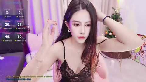 Media: Video of an Asian woman with long black hair, wearing a black lace lingerie top, sitting on a bed, looking at her hand. Background features a Christmas tree and festive decor.