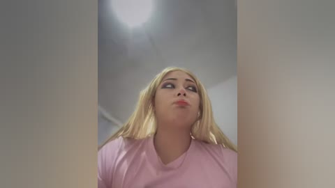 Media: Video of a blonde woman with fair skin, wearing a light pink shirt, gazing upwards at a bright light source, with a soft-focus background.