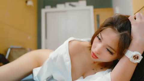 Video of a young Asian woman with long dark hair, fair skin, and red lipstick, lying on a bed in a dimly lit room, wearing a white off-shoulder top, revealing her cleavage, and a wristwatch.