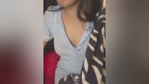 Media: Video of a woman in a light blue, ruffled, unbuttoned dress, revealing cleavage. She has long dark hair and is wearing a zebra print blanket, seated on a red cushioned chair.