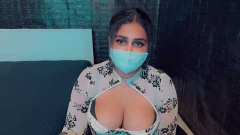 Media: Video of a woman with long dark hair, light skin, and large breasts wearing a white floral-patterned, sheer top revealing cleavage, sitting on a bed with dark curtains and a white headboard.