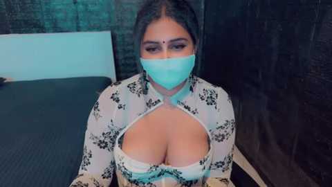 Media: Video of a South Asian woman with medium-dark skin, wearing a turquoise face mask and floral-patterned white blouse, exposing large cleavage, in a dimly lit room with a brick wall.