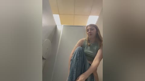 Media: Video of a young woman with long brown hair, wearing a green tank top and blue jeans, crouching in a narrow, dimly lit hallway with fluorescent lighting and white walls.