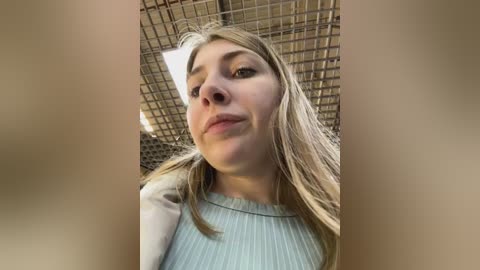 Media: Video of a young woman with straight, shoulder-length blonde hair, wearing a light blue ribbed sweater. She has fair skin and is looking down. Background shows a grid-like metal structure.