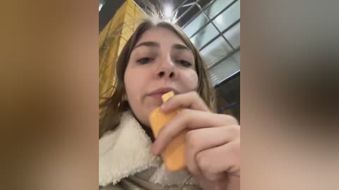 Media: Video of a young woman with long brown hair, wearing a beige jacket, eating a carrot while seated in a dimly lit room with a yellow wall and window blinds.