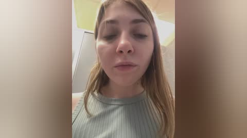 Media: Video of a young woman with fair skin, brown hair, and a light grey ribbed tank top, taken in a bathroom with tiled walls and a yellow ceiling.