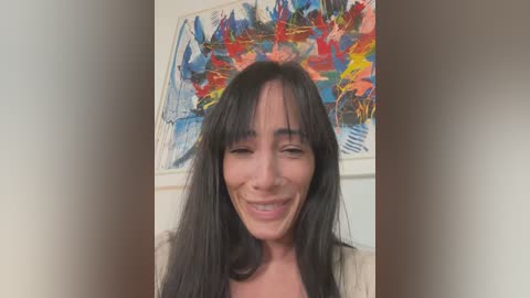 Media: Video of a smiling woman with long black hair, light skin, and bangs, standing in front of a colorful abstract painting on a white wall.