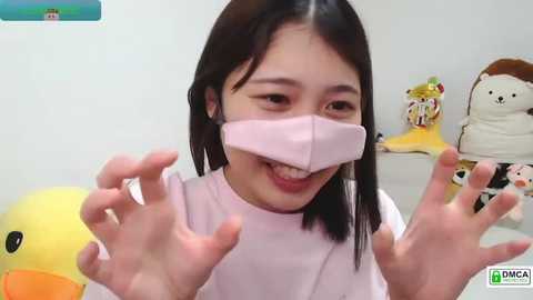 Media: Video of a cheerful Asian woman with dark hair and pink mask, smiling and waving hands, in a child-friendly room with stuffed toys.