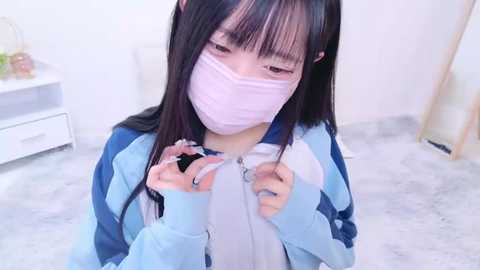 Media: Video of an Asian woman with long black hair, wearing a pink mask, light blue hoodie, and white shirt, adjusting her mask, indoors.