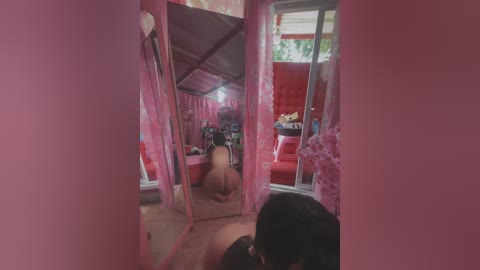 Media: Video of a woman in a pink dress, kneeling on a pink carpet, facing a mirror that reflects a red sofa and a green plant. The room has pink walls and floral curtains.