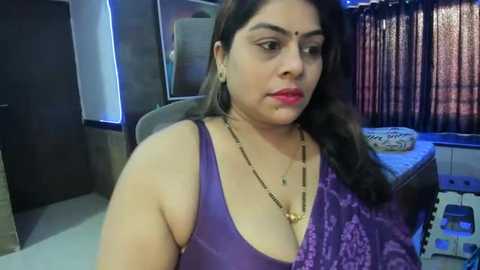 Media: Video of a South Asian woman with medium skin tone, dark hair, wearing a purple sleeveless top, gold necklace, and red lipstick, seated in a dimly lit room with patterned curtains and a white bed.