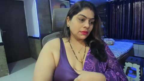 Media: Video of a South Asian woman with medium skin tone and long dark hair, wearing a purple lace blouse, seated at a desk in a modern, dimly-lit bedroom with dark curtains.