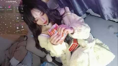 Media: Video of a young Asian woman in a plush white bathrobe, cuddling a teddy bear, sitting on a bed with grey sheets and dark curtains in the background.