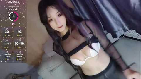 Media: Video of a young Asian woman with long black hair, wearing a black sheer top and white bra, seated on a couch. Virtual reality interface overlay shows health stats and social media notifications.