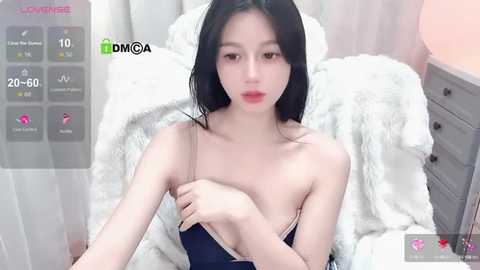 Media: A video of a young Asian woman with long black hair, fair skin, and red lipstick, wearing a dark blue camisole, sitting on a white fur blanket in a bedroom.