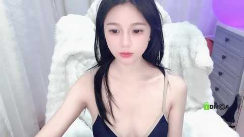 Media: Video of an East Asian woman with long black hair and pale skin, wearing a black tank top with thin straps, sitting on a white bed with a fluffy blanket.