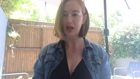 Media: Video of a Caucasian woman with shoulder-length brown hair, wearing a denim jacket over a black top, standing in a sunny backyard with wooden fence and greenery.